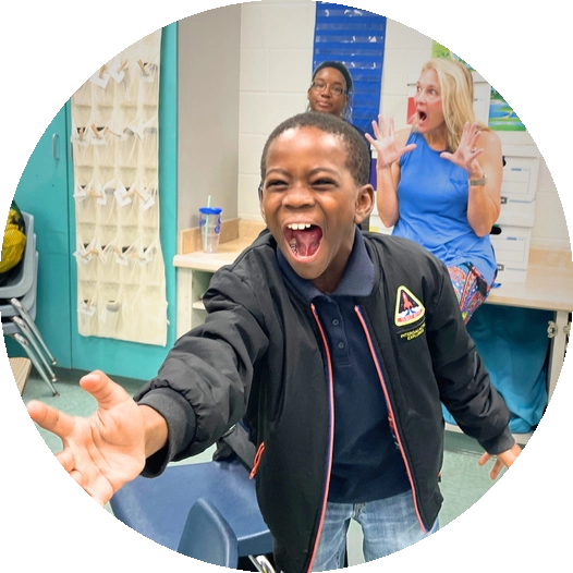 In a vibrant scene of arts-integrated learning, a child in a classroom excitedly gestures with an open mouth and outstretched arm. A woman in a blue dress reacts dramatically from her perch on a desk, while another adult watches from behind with an approving smile.