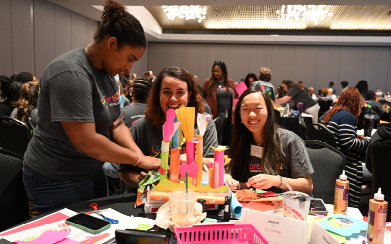 Three people work together on a colorful arts and crafts project at a table, smiling and engaged. They are in a large room with others, and supplies like glue and paper are spread out. The atmosphere is lively and creative.