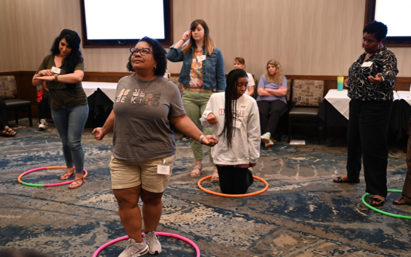 A group of people is engaged in an indoor activity. Some are standing, while others kneel inside colorful hula hoops. One woman wears a "BE KIND" shirt. Tables and chairs are visible in the background.