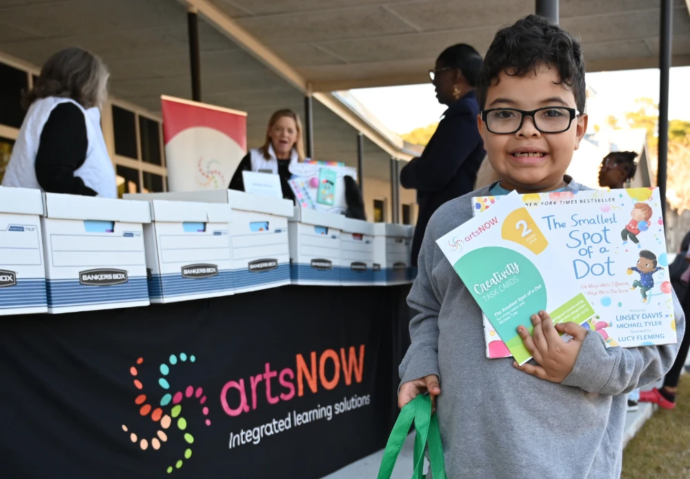 A smiling child holds up books in front of a table with an artsNOW banner, while adults, including SC Arts Alliance’s Jeff Robinson, distribute materials and promote arts advocacy in the background. A smiling child holds up books in front of a table with an artsNOW banner, while adults, including SC Arts Alliance’s Jeff Robinson, distribute materials and promote arts advocacy in the background.