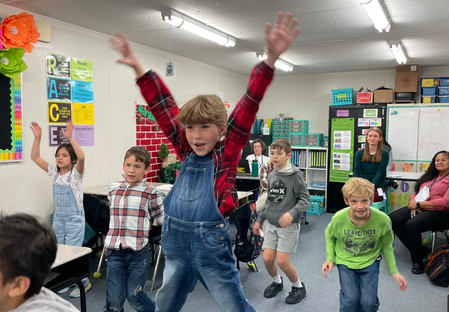 Children in a classroom participate in an energetic summer activity; one child in denim overalls jumps with arms raised while other students creating and smiling move around. Teachers and students are visible in the background.
