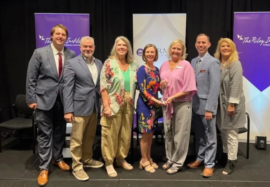 Seven adults stand together on a stage, smiling at the camera. Dressed in business attire, they stand before banners for "The Riley Institute," celebrating a federal grant supporting ArtsNOW’s arts-integrated STEM learning initiatives.