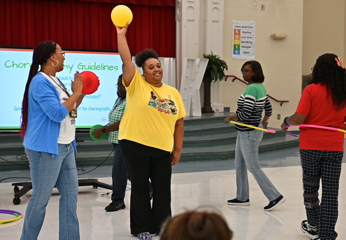 Four women stand in a gymnasium for One-Day Events, each holding colorful props. One woman in a yellow shirt raises a yellow ball, smiling. A choreographed activity is underway, with a guidelines screen visible in the background.