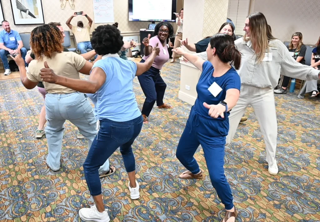 A group of people enthusiastically dance in a circle in a carpeted conference room, smiling and laughing, embracing learning and joy in a changing world, while others sit and watch in the background.