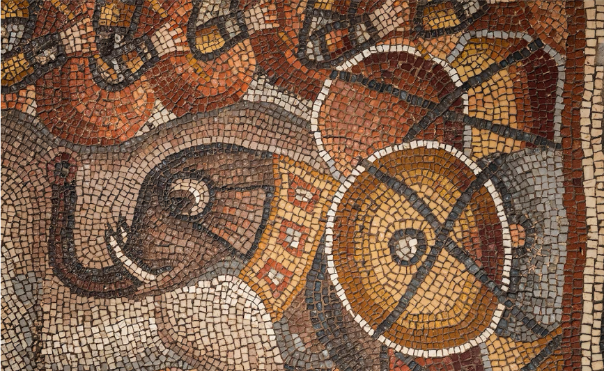 Ancient math mosaic depicting an elephant adorned with decorative patterns, standing among round shields featuring geometric designs in shades of brown, yellow, and red—a striking tribute to division and multiplication in art.