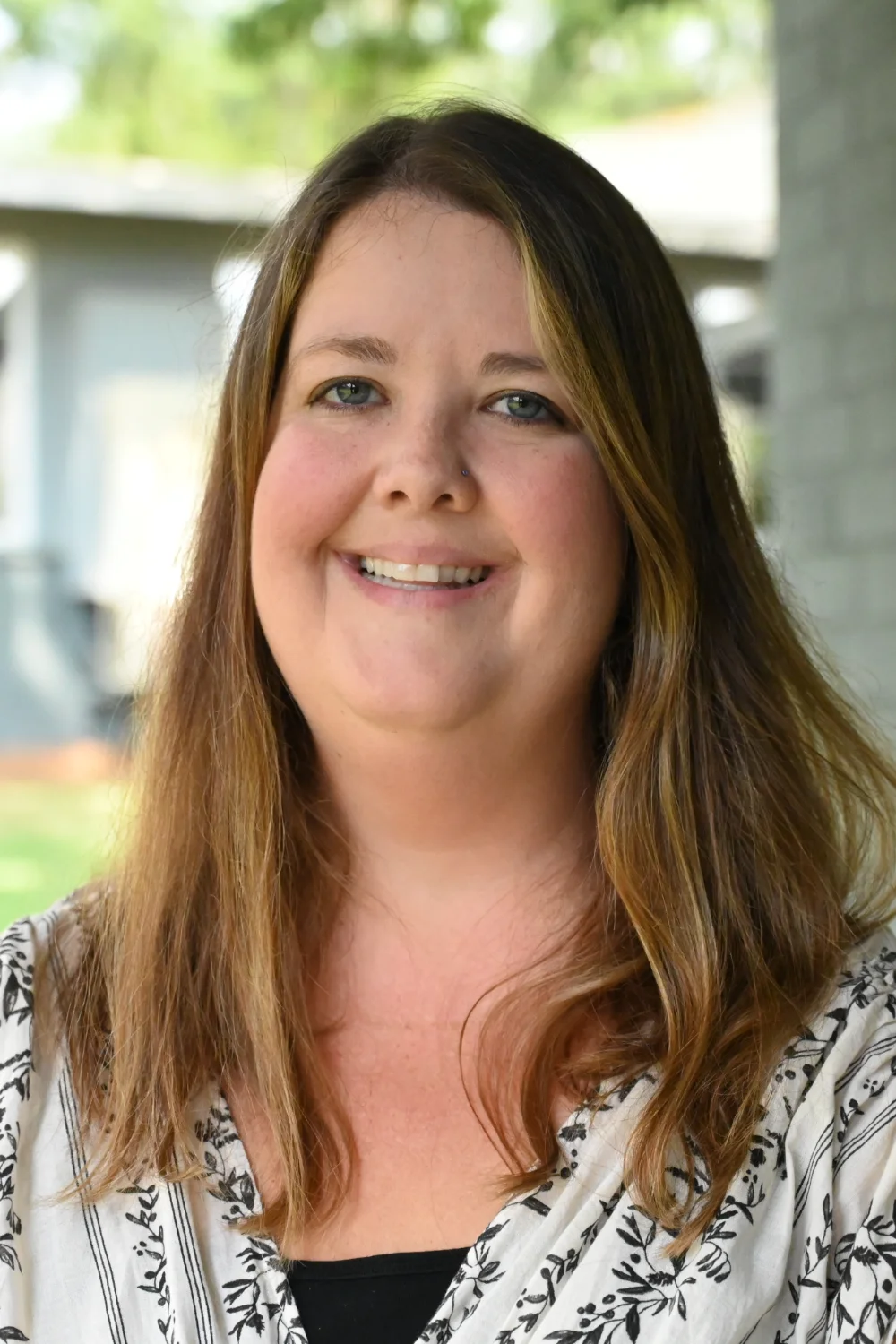 Stephanie Lancaster, a woman with long brown hair, wearing a patterned white and black blouse, smiles at the camera outdoors with greenery and a building blurred in the background.